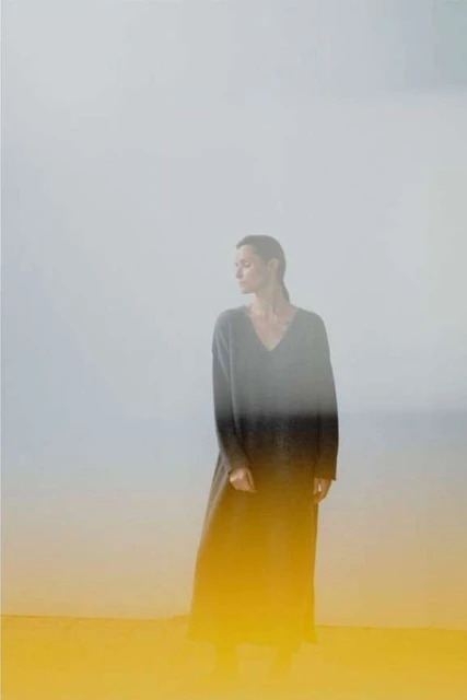 A woman in a long-sleeve maxi dress poses against a foggy grey background with yellow tint, looking off to the side in a fashion editorial.