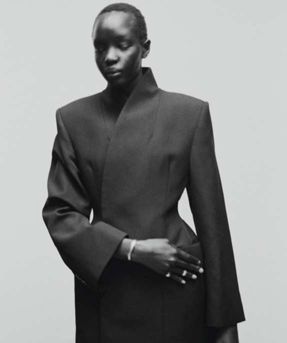 Elegant studio shot of a black model in a chic, dark jacket with sharp lines, set against a neutral background. Classic fashion portrait.