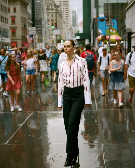 Fashionable woman walks on a wet city sidewalk, surrounded by blurred pedestrians and buildings in the background, captured in medium shot.