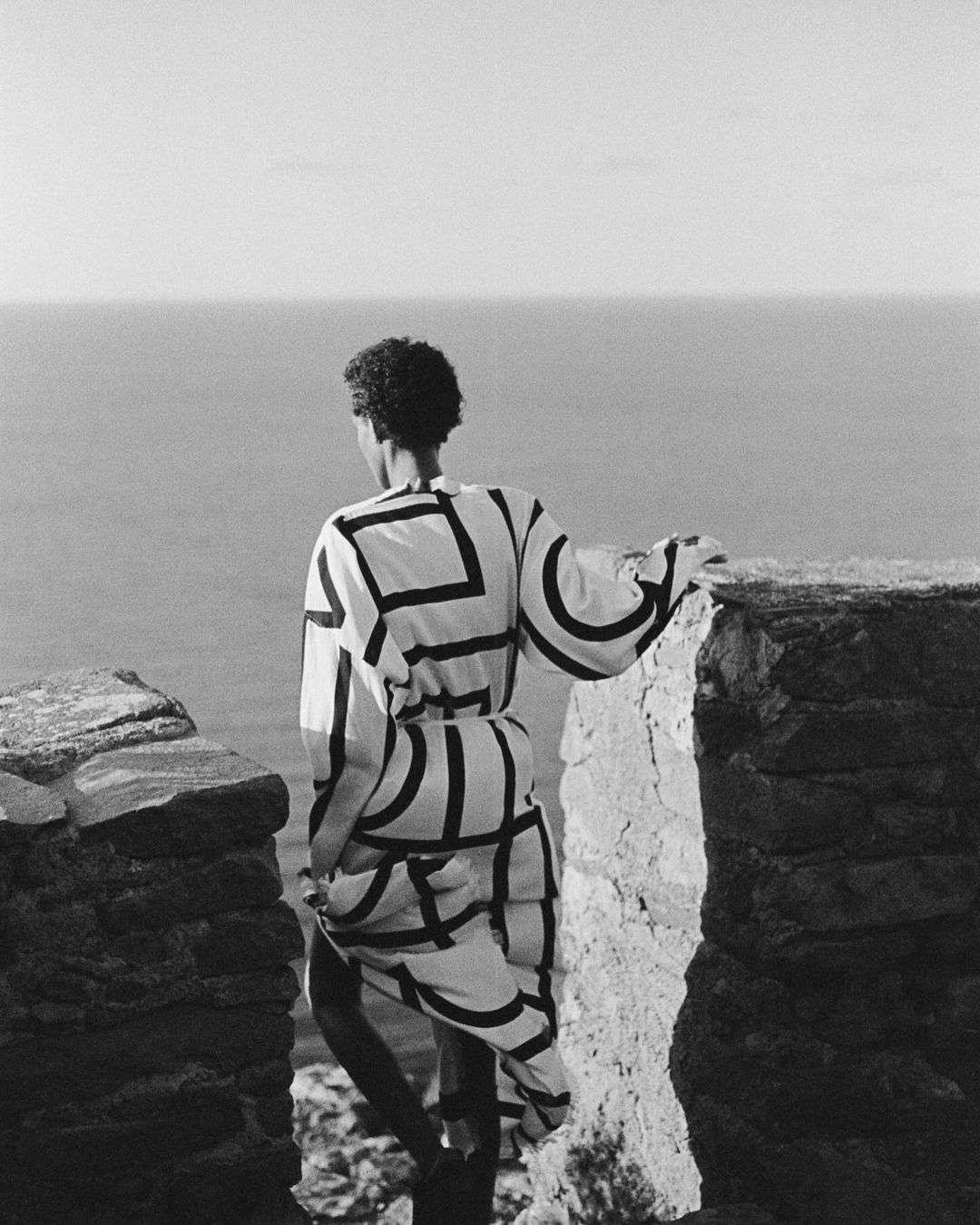 Monochrome fashion shot of an adult female with curly hair, standing on a stone wall in a dress with a geometric pattern looking at the ocean.