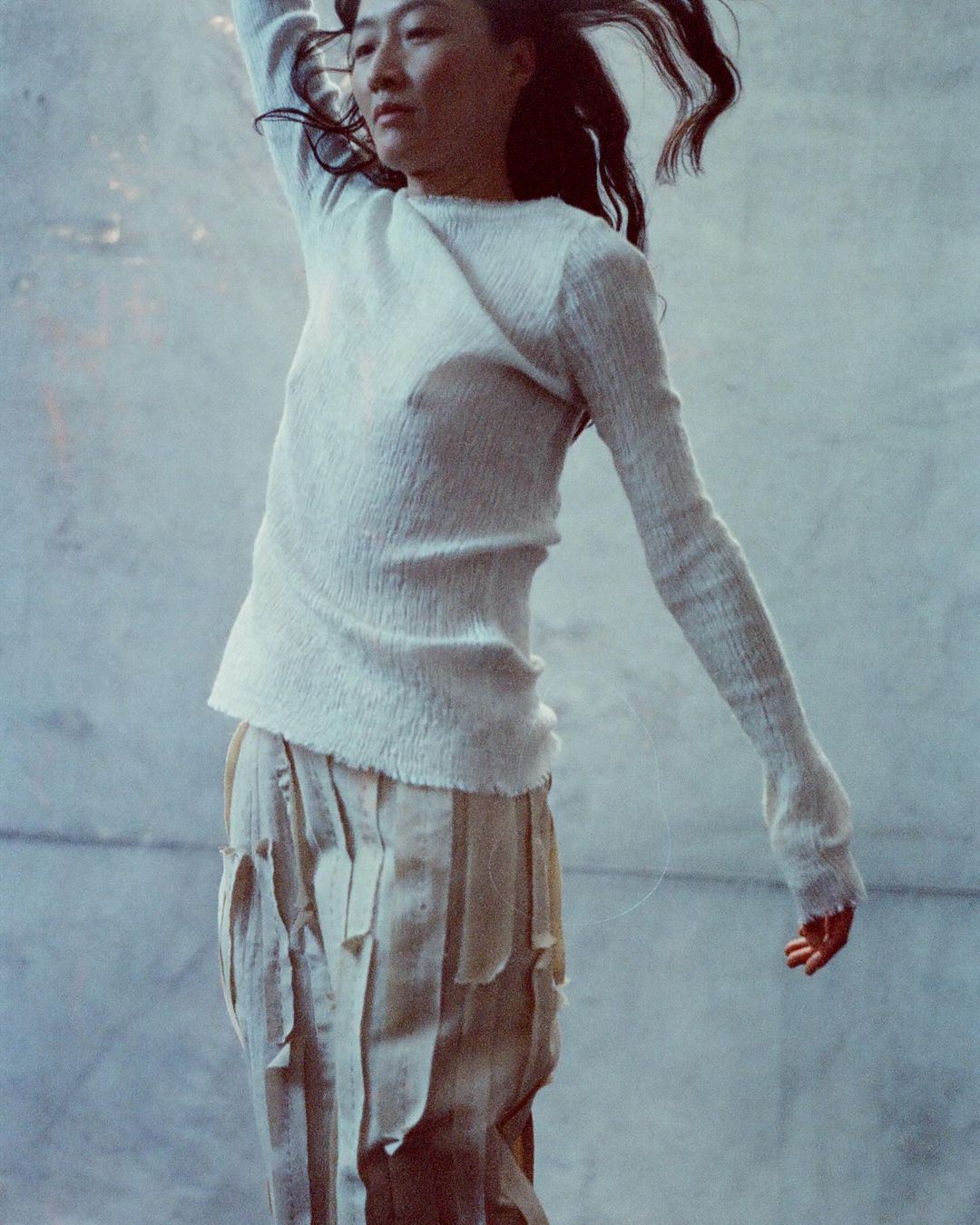 Asian woman in motion, wearing an off-white sweater and light-colored textured skirt, against a light grey, textured background.