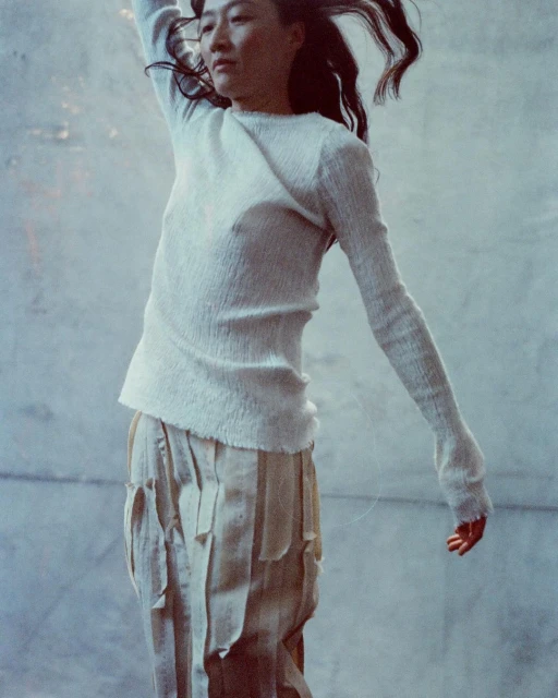 Asian woman in motion, wearing an off-white sweater and light-colored textured skirt, against a light grey, textured background.