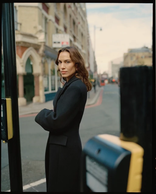 A woman in a black coat stands on a city street in London, with buildings in the background and a pedestrian crossing signal in the foreground.