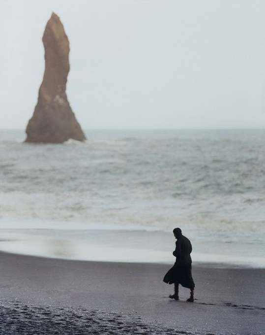 A person in a dark coat walks on a black sand beach towards a sea stack under an overcast sky, creating a moody and atmospheric scene.