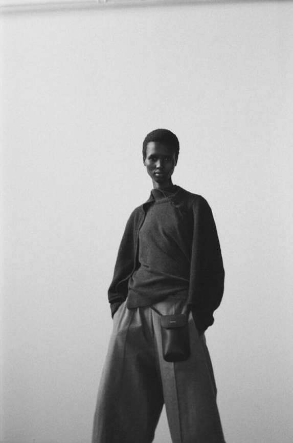 Monochrome portrait of an African woman wearing a dark top and light pants with a small bag against a neutral backdrop.