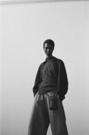 Monochrome portrait of an African woman wearing a dark top and light pants with a small bag against a neutral backdrop.