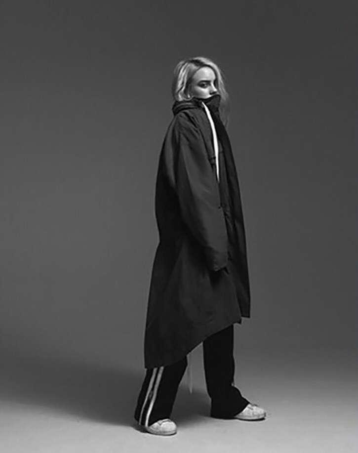 Portrait of Billie Eilish wearing an oversized dark jacket and striped track pants against a grey seamless studio backdrop.