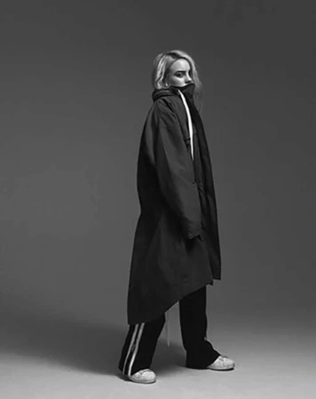 Portrait of Billie Eilish wearing an oversized dark jacket and striped track pants against a grey seamless studio backdrop.