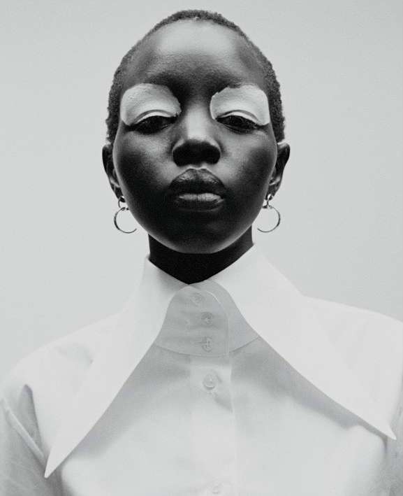 Striking portrait of a dark-skinned woman with shaved head, closed eyes with white eyeshadow, wearing a white collared shirt against a neutral background.