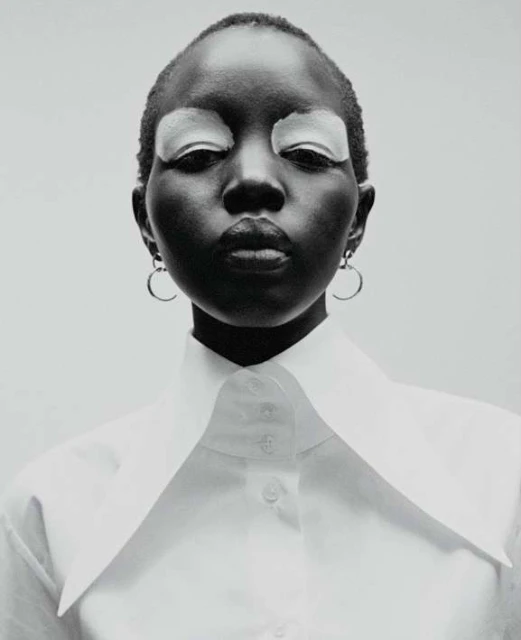 Striking portrait of a dark-skinned woman with shaved head, closed eyes with white eyeshadow, wearing a white collared shirt against a neutral background.