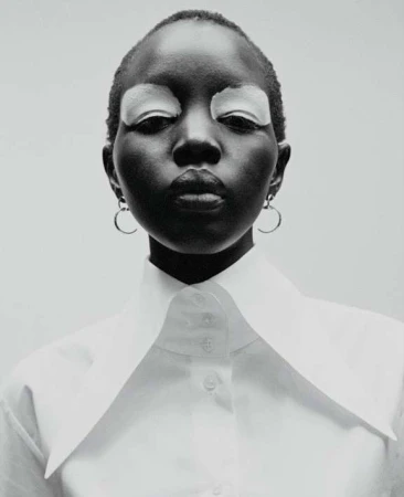 Striking portrait of a dark-skinned woman with shaved head, closed eyes with white eyeshadow, wearing a white collared shirt against a neutral background.