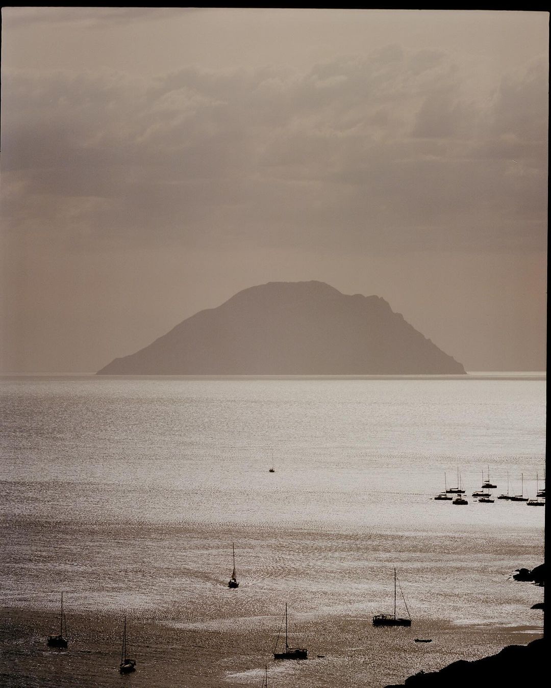 Overcast seascape view of sailboats on the water with an island in the distance under a cloudy sky, creating a peaceful, muted scene.