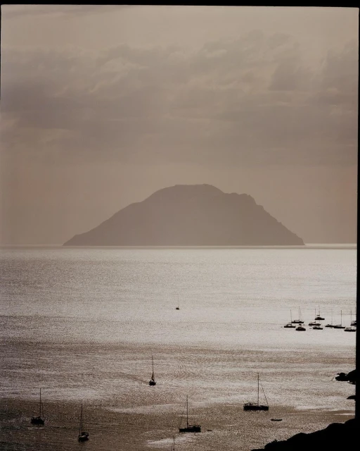 Overcast seascape view of sailboats on the water with an island in the distance under a cloudy sky, creating a peaceful, muted scene.