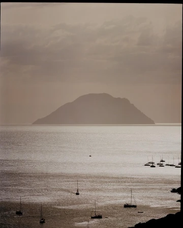 Overcast seascape view of sailboats on the water with an island in the distance under a cloudy sky, creating a peaceful, muted scene.