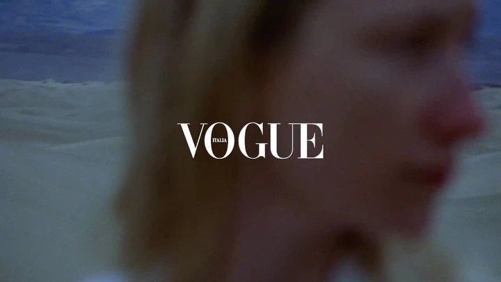 Fashion editorial shot with a blurred female face and Vogue Italia logo against a blurred desert dune landscape under a dusk sky.