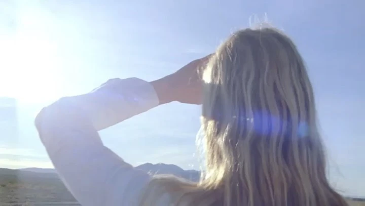 Adult female shields her eyes while gazing across a desert landscape under a sunny sky; seen from a medium shot showing her blonde hair.