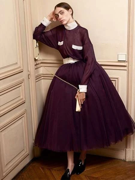 Adult woman poses near a door in a sheer plum dress, white belt, and black shoes in a fashion photography studio shoot.