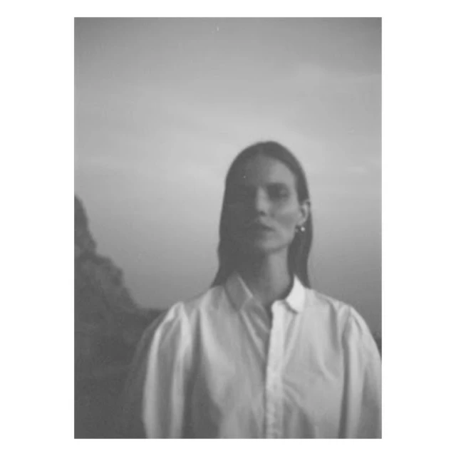 Black and white portrait of a woman in a button-down shirt, set against a neutral sky and shadowed natural landscape backdrop.