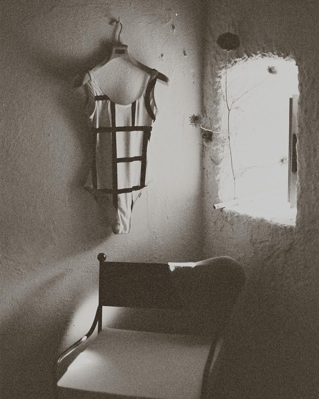 Black and white photo of a bathing suit hanging on a wall next to a window, with a chair in the foreground of an indoor setting.
