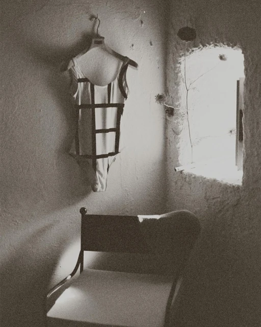 Black and white photo of a bathing suit hanging on a wall next to a window, with a chair in the foreground of an indoor setting.