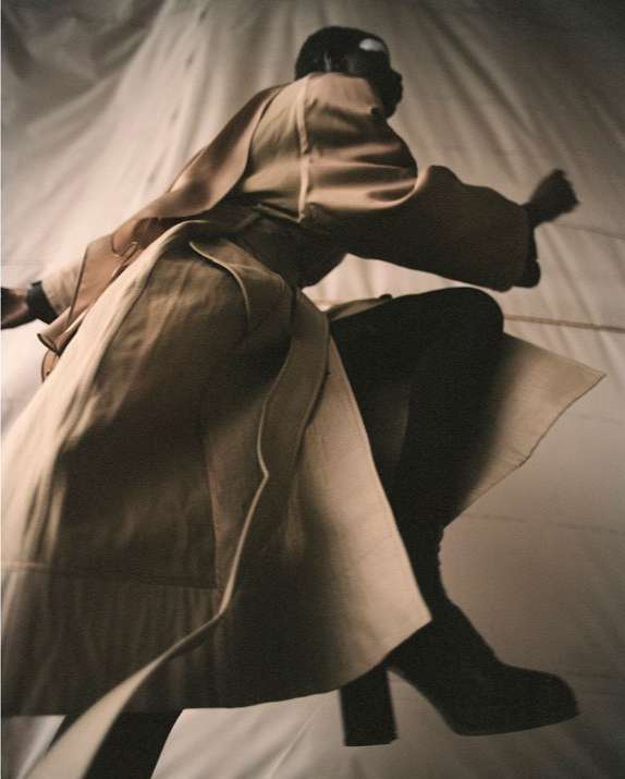 An adult female fashion model with dark skin in a long trench coat leaps in the air against a white background in a studio shot.