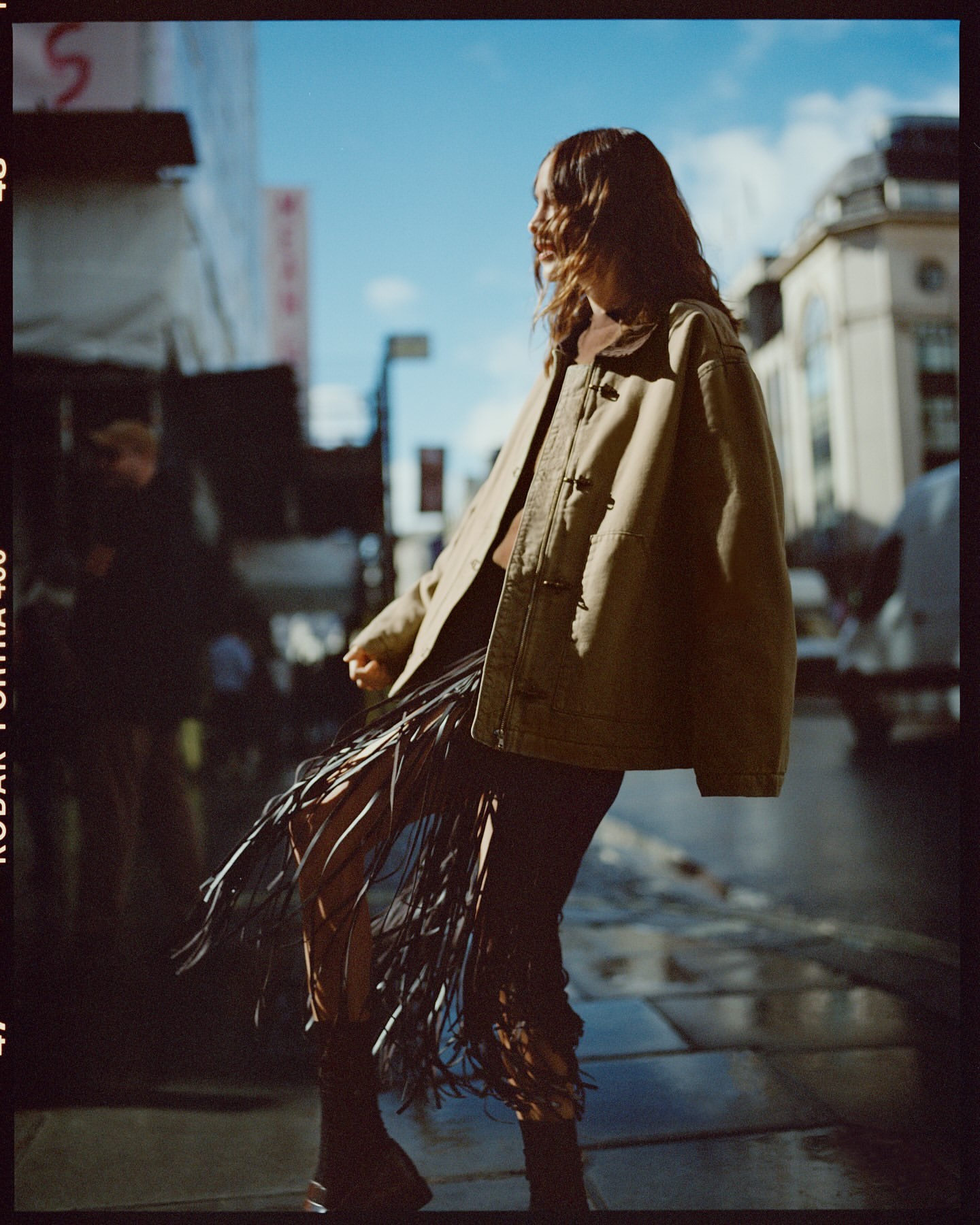 A woman in a tan jacket and fringe skirt stands on a city sidewalk on a sunny day, captured in a fashion-editorial style photograph.