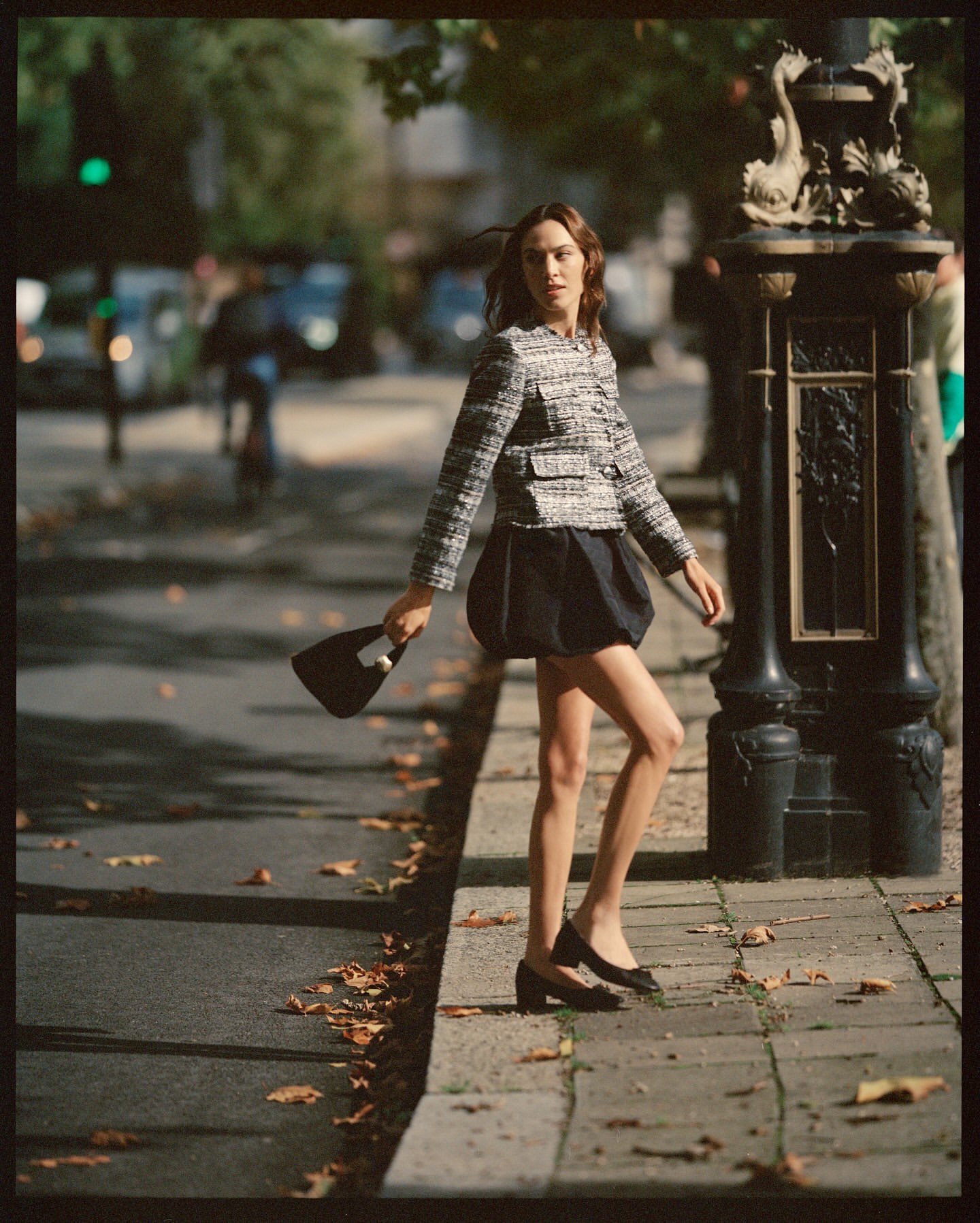A stylish woman in a tweed jacket and dark skirt walks along a city sidewalk holding a purse, on a sunny day with tree shadows.
