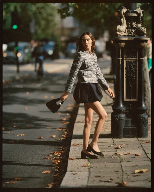 A stylish woman in a tweed jacket and dark skirt walks along a city sidewalk holding a purse, on a sunny day with tree shadows.