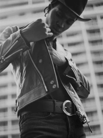 Black and white portrait of a man in New York wearing a leather jacket and cowboy hat adjusting his jacket.