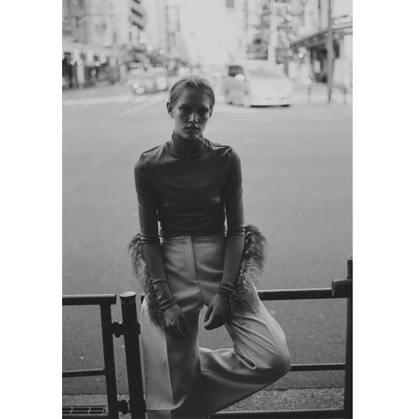 Monochrome outdoor fashion shot of a woman in a turtleneck and wide-leg pants, leaning against a railing on a city street.