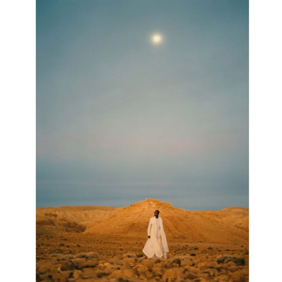 A single Black adult stands in a light-colored coat in a desert landscape under a bright sky with a bright light source above.