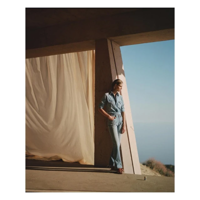Woman in denim leans against a concrete structure with curtain, set against a backdrop of sea and sky on a sunny day.