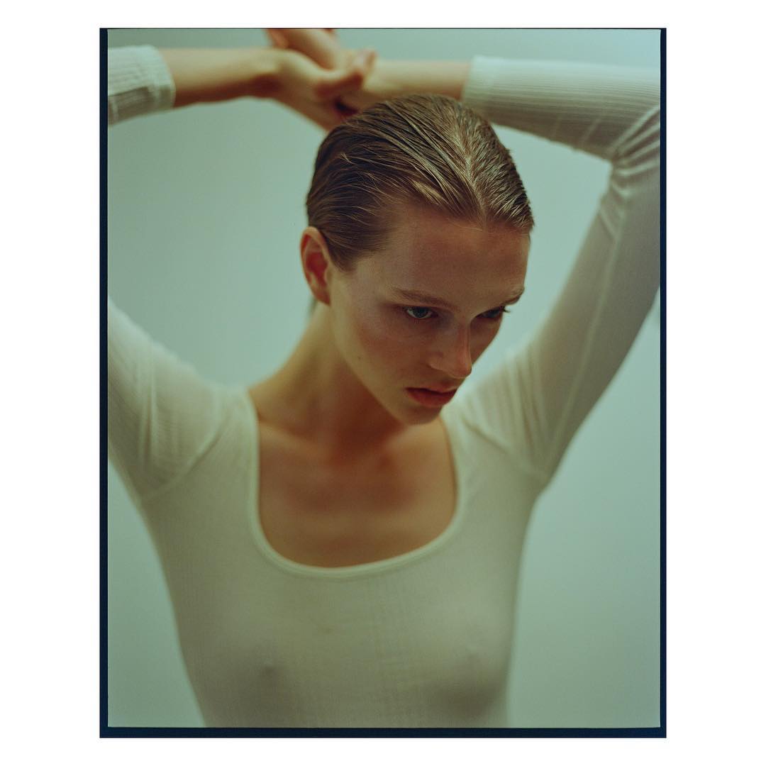 Portrait of a young adult woman with wet slicked-back hair, wearing a long-sleeved white top against a pale greenish background, eye-level shot.