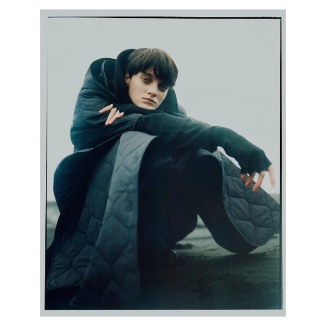 A person in dark clothing, wearing a quilted coat with a hood, sits in a relaxed pose against a light grey backdrop in a studio shot.