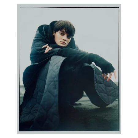 A person in dark clothing, wearing a quilted coat with a hood, sits in a relaxed pose against a light grey backdrop in a studio shot.