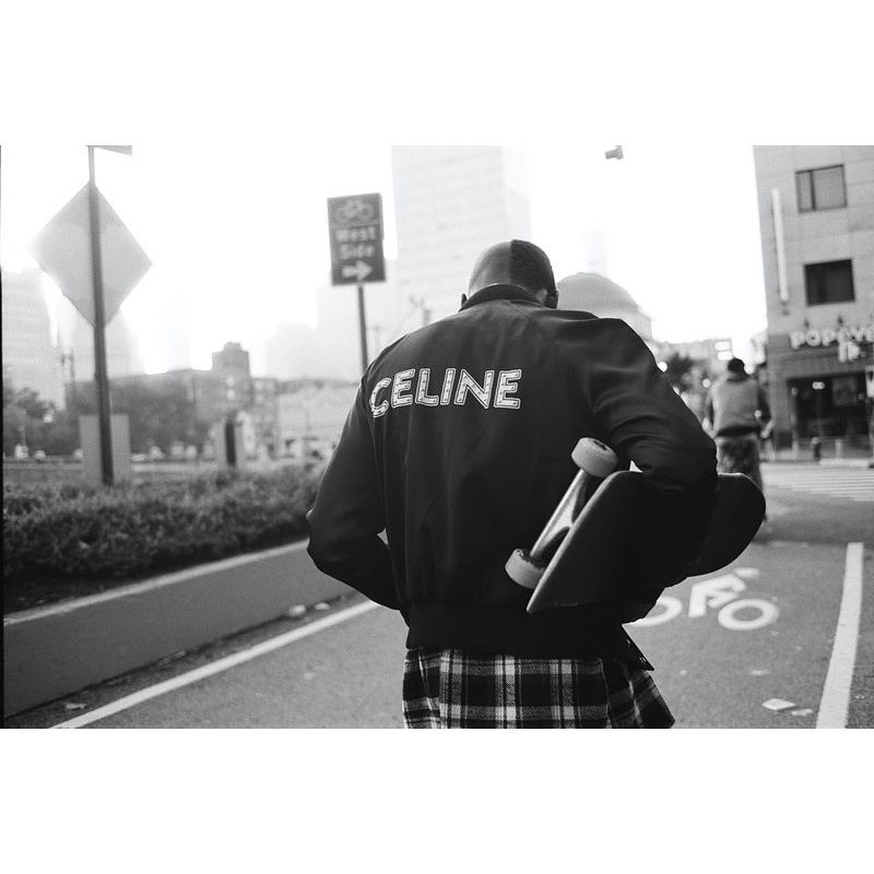 Person with skateboard walks on city street. Figure wears a black bomber jacket with 'CELINE' printed on the back, and a plaid skirt.