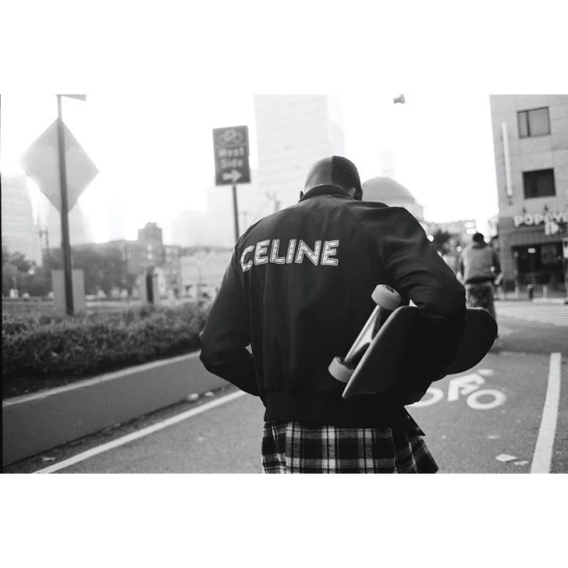 Person with skateboard walks on city street. Figure wears a black bomber jacket with 'CELINE' printed on the back, and a plaid skirt.