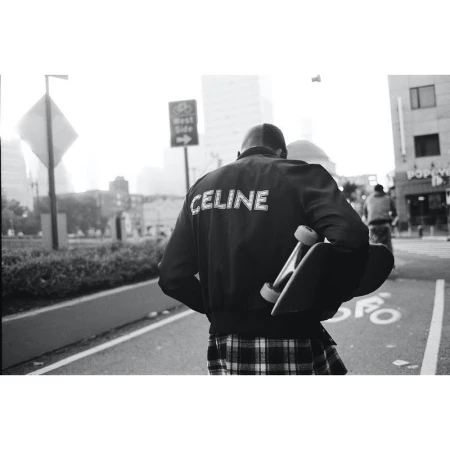 Person with skateboard walks on city street. Figure wears a black bomber jacket with 'CELINE' printed on the back, and a plaid skirt.