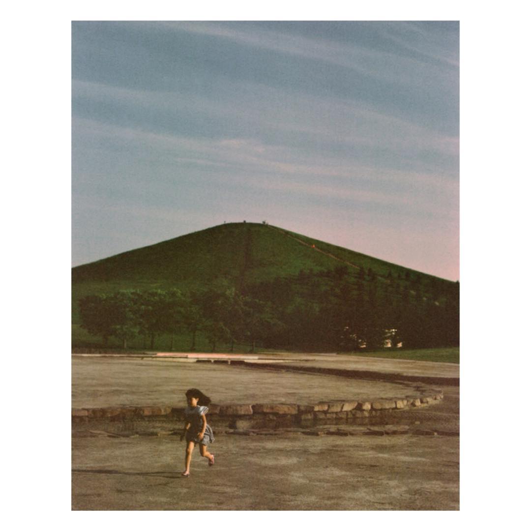 Girl running barefoot in a park toward a large green hill under a partly cloudy sky, captured in a landscape orientation.