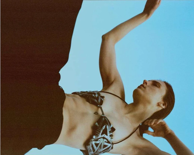 A woman in a geometric print top poses in a low angle shot against a light blue sky, with dark pants visible on the left side.