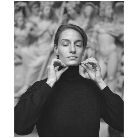 Monochrome portrait of a woman in a dark turtleneck sweater, touching her neck, with blurred statues in the background.