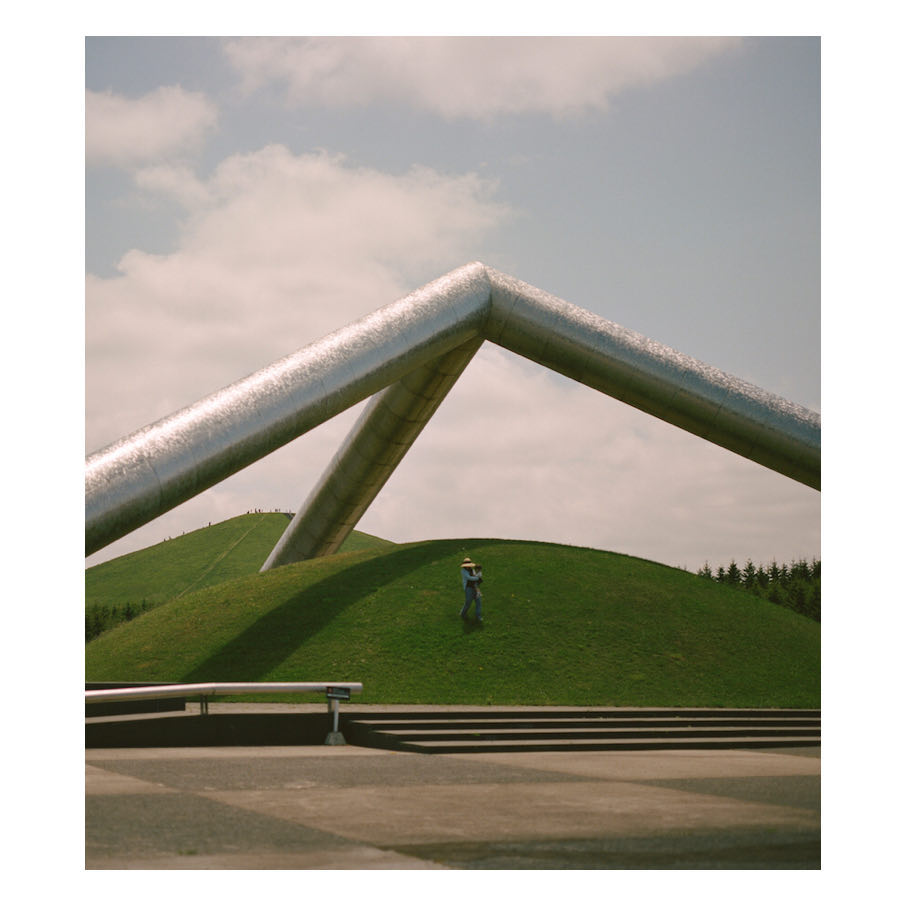 Figure standing on a green hill under a metallic A-frame structure, set against a partly cloudy sky. Steps and a bench in the foreground.