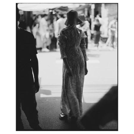 Monochrome street scene featuring a person in a dress and hat, viewed from behind, walking into a crowded area in soft, diffused light.