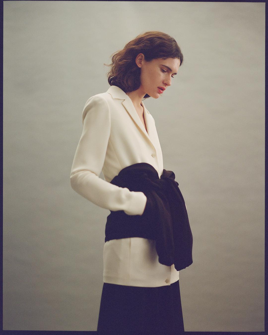 Elegant woman in a cream blazer and black skirt gazes downward, holding a dark garment in a minimalist, indoor studio setting.
