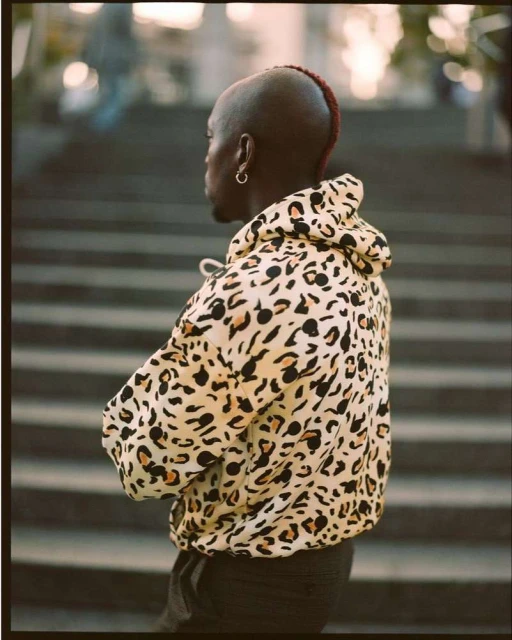 An adult male stands in a stairwell with his arms crossed, wearing a hooded sweater with a leopard print design, side profile view.