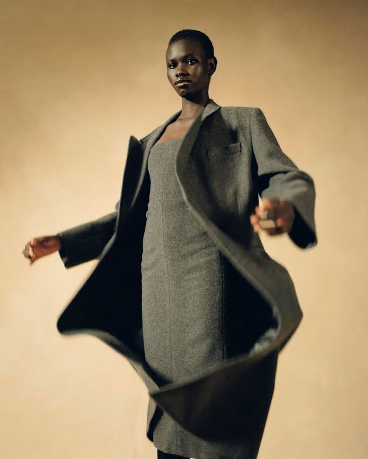 Fashion shot of a Black female adult model wearing a gray wool dress and matching overcoat in a studio setting, mid-motion.