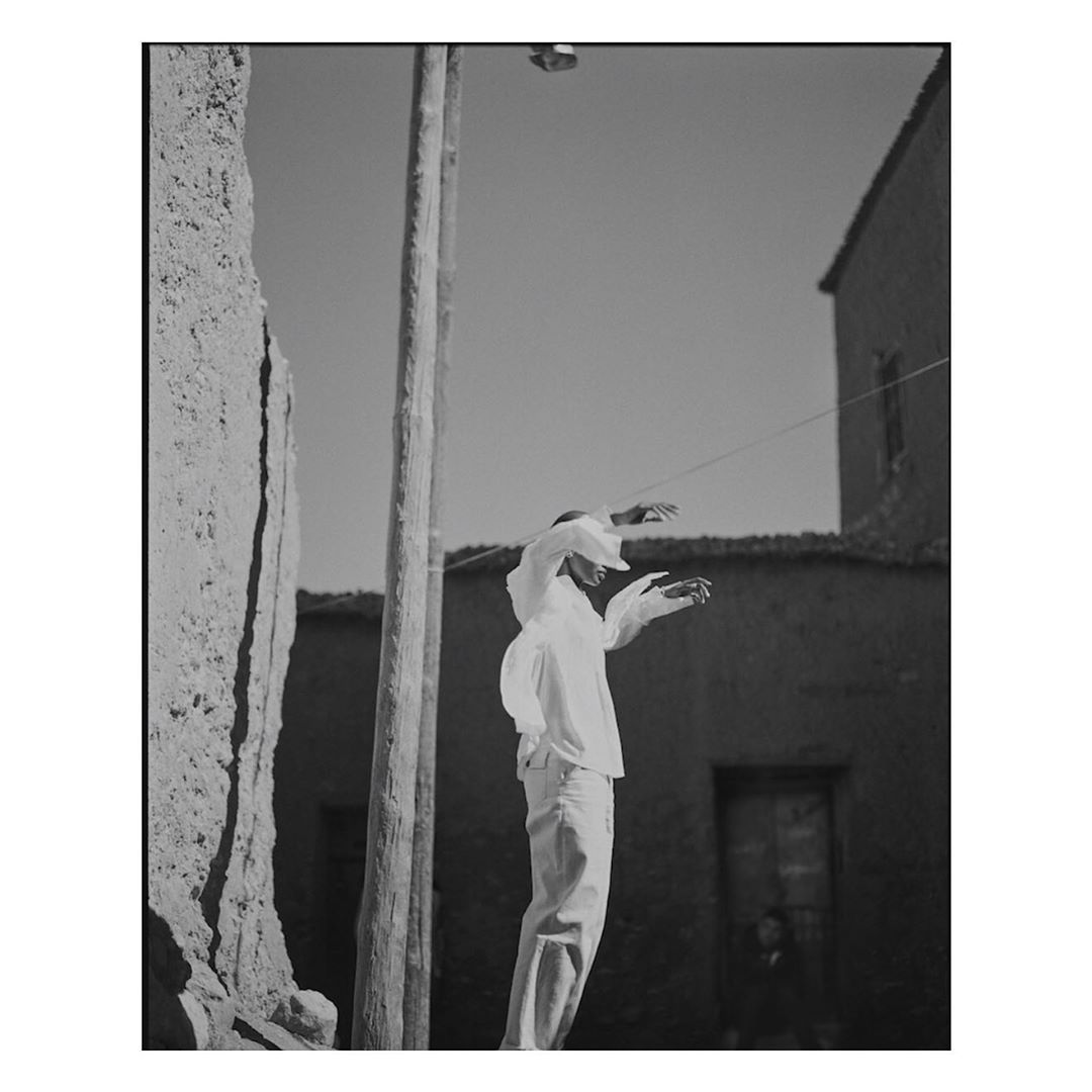 Monochrome image of an adult wearing white clothing standing outdoors with arms raised near weathered buildings and a utility pole.