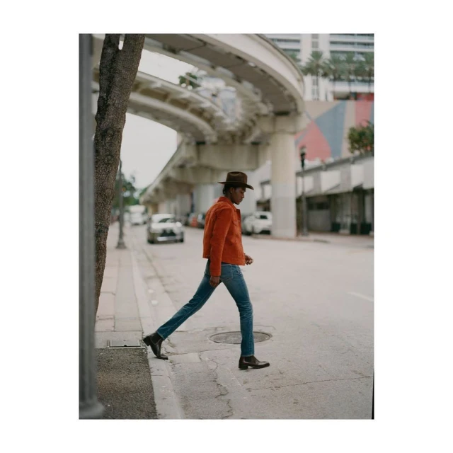 An adult strides across a crosswalk in jeans and a rust colored jacket, with an elevated transit line curving above the city street.