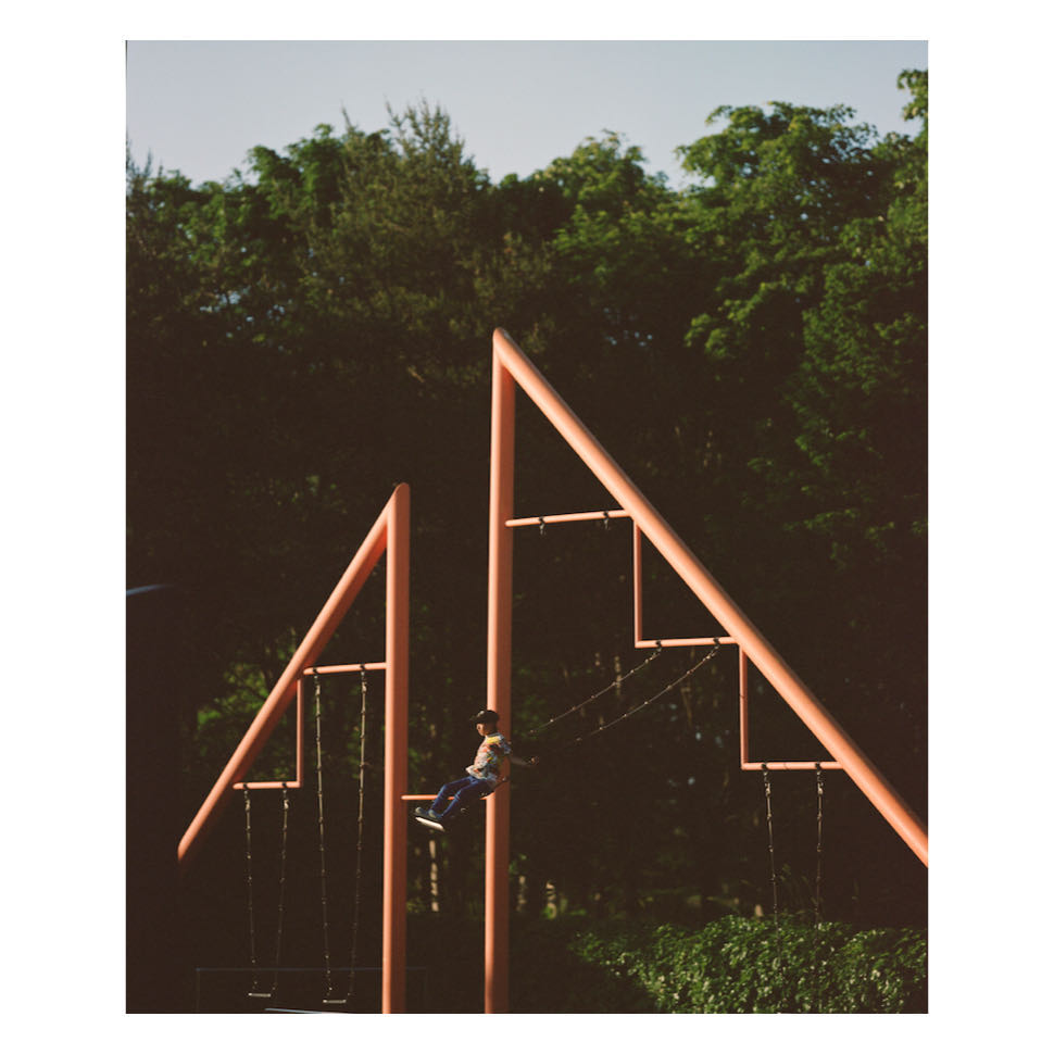 Child swings on a pink metal swing set, surrounded by lush green trees under a bright sky. Outdoor playground in a park setting.