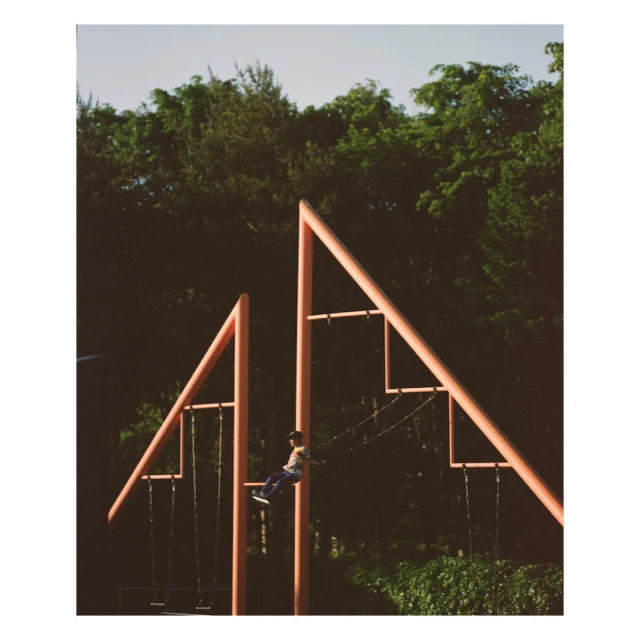 Child swings on a pink metal swing set, surrounded by lush green trees under a bright sky. Outdoor playground in a park setting.
